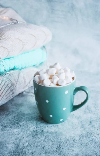 Turquoise cup, mug of hot drink with white marshmallow on the background of stack of winter knitted clothes. Winter cozy still life. Hot beverage, cacao, milk, coffee.