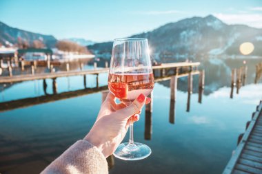 Female hand with glass of rose wine. Cozy pier on the coast of the lake Tegernsee. Alpine mountains in Bavaria. Mountain view, beautiful landscape in Germany. Adventure in Europe.