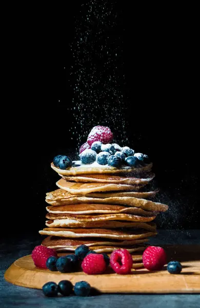 Stack of pancakes with blueberry, raspberries and honey on the wooden cutting board. Breakfast, food photo. Dark background.