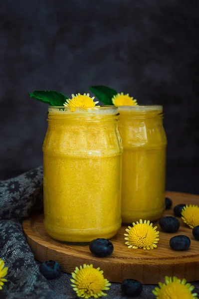 Yellow mango smoothies with flowers, berries, wooden plate on a dark background. Food and drink photography. Healthy vegan recipe
