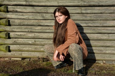 Young girl crouches on the floor in front of old wooden planks in the sun  and looks forward