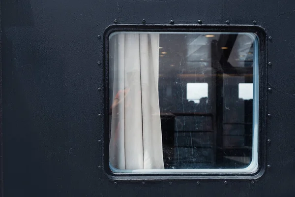 Window view on the ferry. Metallic, dark blue colored ferry wall with screws around the window. White curtains inside can be seen through the glass. Abstract, minimalistic picture taken in a horizontal position.