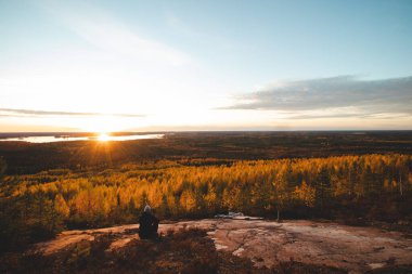 Finlandiya, Paltamo yakınlarındaki bir kayadan nefes kesici bir gün batımı. Bir maceracı bir kayanın üzerinde dinlenir ve bir turuncu kürenin ağaçların renkli yapraklarını aydınlatmasını izler. Sonbahar renkleri.