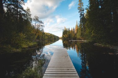 View of a wooden pier and a still river reflecting the colourful autumn forest with all its colours. Wonderful autumn in the barren wilderness of Hossa National Park, in Lapland, northern Finland.