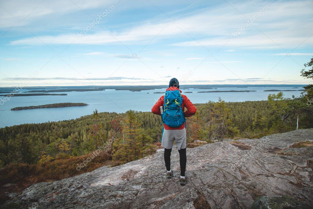 hiker wearing a jacket and carrying a backpack, standing on a rock watching Lake Jatkonjarvi at sunset in Koli National Park, eastern Finland. A man aged 24 wearing sports clothes. Active lifestyle.
