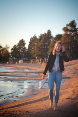 Gülümseyen genç esmer sporcu, kumlu bir sahilde kot pantolon ve siyah bir bluzla çıplak ayakla günbatımının ve son ılık ışıkların tadını çıkarıyor. Sotkamo, Finlandiya. İskandinavya 'yı keşfetmek. Gerçek insanlar..