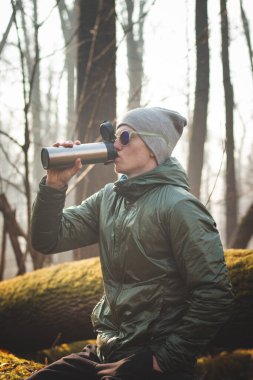 Yabanda bir ağaç gövdesinde oturan tecrübeli bir yürüyüşçü yürüyüş sırasında termostan sıcak çayını içiyor. Avrupa ormanlarının biyolojik çeşitliliğini keşfeden gerçek insanlar. Gerçek gülümseme.