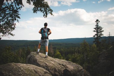 Swarthier 'in tipi bir kayanın tepesinde dikilmiş Zdarske Vrchy' deki bir vadiye bakıyor Çek Cumhuriyeti 'ndeki bir dağ sırasına. Sırt çantalı adam zorlu bir tırmanıştan hoşlanıyor..
