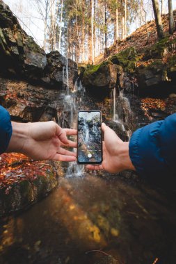 Tourist takes a picture of Tosanovsky waterfall on his mobile phone for his social networks and for his memories. Beskydy mountains, Czech republic, centre of Europe.