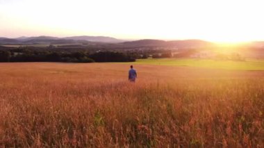 Thoughtful man in a colourful shirt walks in the early evening light through a meadow teeming with life and biodiversity, wondering how to preserve this healthy ecosystem.