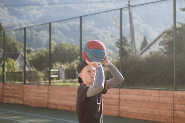 Spor giyim antrenmanındaki sarışın çocuk üç sayılık çizginin arkasından basket atıyor. Açık hava basketbol sahası. Mevsime hazırlanıyorum..