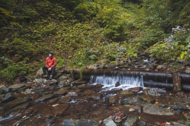 Kırmızı ceketli aktif bir yürüyüşçü zorlu bir patikadan sonra bir dağ nehrinin yanında oturur ve cam bir konteynırdan temiz su içerek dinlenir. Vahşi yaşam.