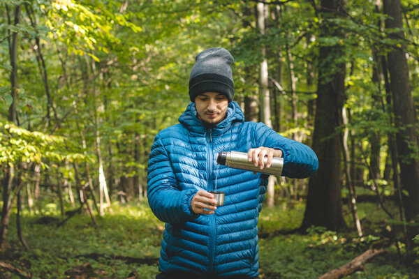 Hiker on a long trail takes a rest and pours hot tea from a thermos into a mug to warm up and replenish vitamins and fluids. High mountain hiking.