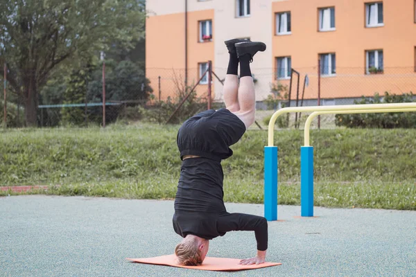 Calisthenics athlete performs a headstand using a solid core and arm stability on an orange mat on an outdoor workout court. The middle phase of the exercise.