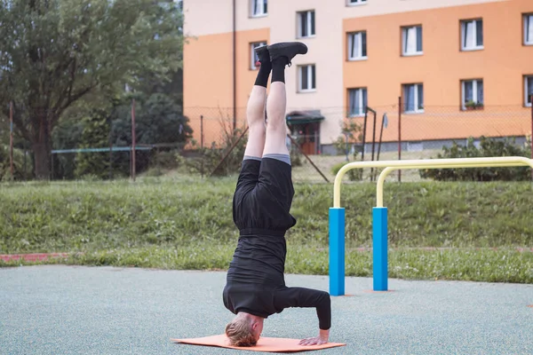 Calisthenics athlete performs a headstand using a solid core and arm stability on an orange mat on an outdoor workout court. The final phase of the exercise.