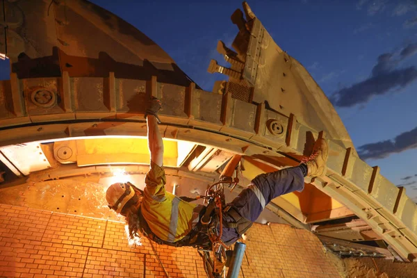 Rope access maintenance welder abseiler wearing fall safety body harness helmet protection hanging upside way welding repairing reclaimer wheels bucket construction mine site Perth, Australia 