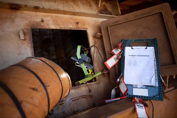 Safety workplace permit to work paper clipping on isolation control permit board together with red personal danger locks and tags at confined space entry exit door ventilation fan while worker is working inside                  
