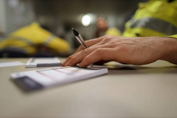 Siyah kalem tutan ve elini kişisel güvenlik risk değerlendirmesinin üzerine koyan yüksek riskli işçiler, arka planda çözülmüş takım çalışmasıyla birlikte masaya beş broşür koyuyorlar.  