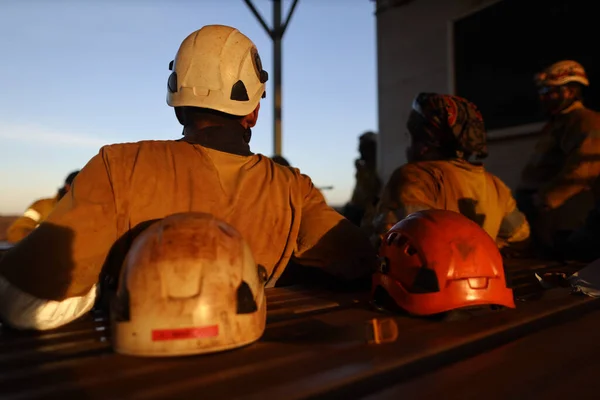 Rope access miner setting having a rest during extreme hot weather in Pilbara WA. wearing white safety side impact helmet head fall protection with defocused co workers setting beside and at the background construction mine site Perth
