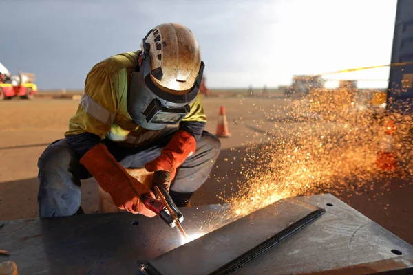 Trained construction miner welder boilermaker worker setting wearing a safety helmet, red welding leather glove dark face shield protection while commencing hot work gouging metal plate on the ground surface construction 