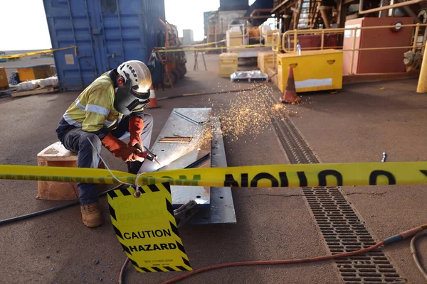 Safety work practices worker setting in comfortable position wearing a safety helmet, red welding safety leather glove, dark face shield protection using yellow striped caution tape warning sign barricade exclusion zone preventing from public access 