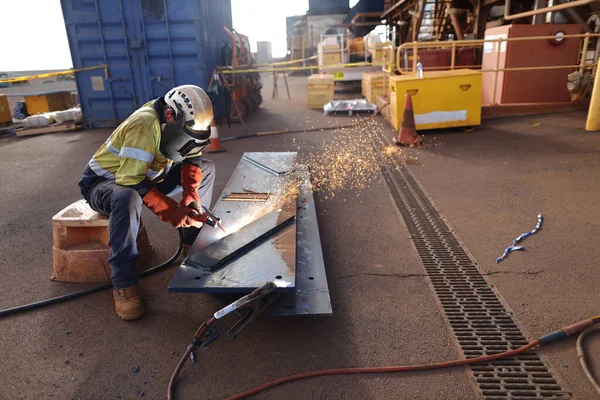 Safety work practices worker setting in comfortable position wearing a safety helmet, red welding safety leather glove, dark face shield protection while commencing hot work gouging metal plate on the ground surface construction mine site   