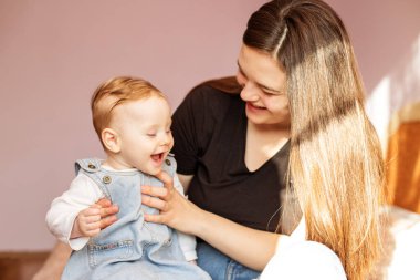Happy mom holds little daughter on lap. Mother and little todler girl are playing in room. Denim sundress. Bright sunny light.