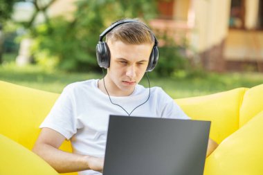 Beyaz tişörtlü meşgul adam sarı fasulye koltukta otururken dizüstü bilgisayarla çalışıyor..