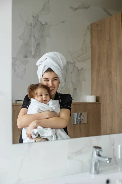 Mom and baby look in mirror and smile in bathroom. White towels. Cozy house. Family time.