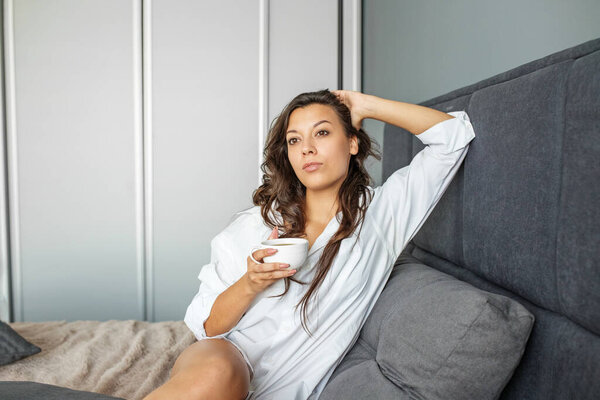 Girl with coffee. Home comfort. Mental health. Woman in a white shirt in bed. Modern bedroom interior in gray. pleasant weekend getaway at home.