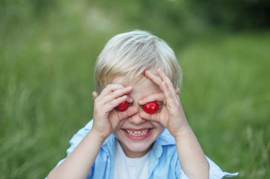 Şirin sarışın çocuk, dışarıda kiraz yiyor, komik suratlar yapıyor ve kirazla oynuyor, eğleniyor.