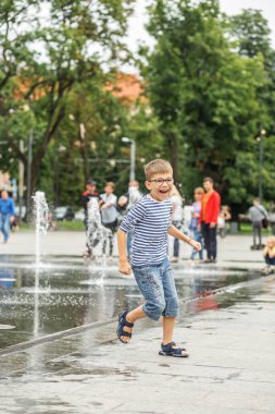 Küçük mutlu çocuk yaz günü kuru çeşmedeki su fıskiyeleri arasında kare şeklinde oynuyor. Şehirdeki çocuklar için aktif bir yaz tatili. Çizgili gömlek ve kot şortlu çocuk..