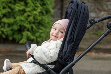 Little very beautiful girl is sitting in stylish black stroller. Gently pink hat and beige jacket. Spring and summer season.