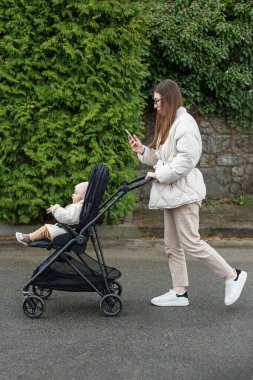 Doğum izninde zaman geçirmek. Anne bebek arabasında çocuk taşıyor ve mobil cihaz kullanıyor..