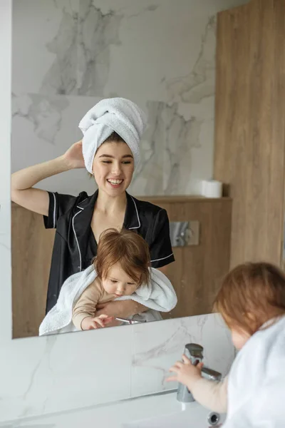 Mom and baby look in mirror and smile in bathroom. White towels. Cozy house. Family time.