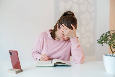 Ev ofisi, uzaktan çalışma, e-öğrenme konsepti. Kız not defterine not alıyor. Cep telefonu standı.