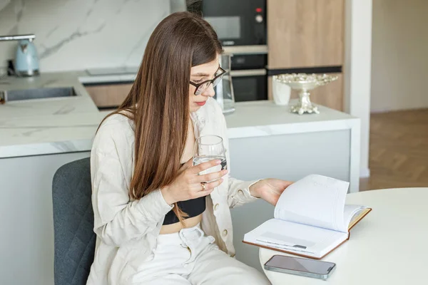 若い女性は台所のテーブルに座って水を飲んで日記を通して見ている 一日計画を立てる — ストック写真