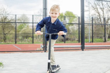 Küçük sarışın çocuk scooter kullanıyor. Çocuk güneşli yaz gününde spor sahasında oynuyor. Sağlıklı açık hava aktiviteleri. Okula giden tatlı bir okul çocuğu..