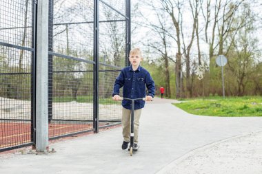 Küçük sarışın çocuk scooter kullanıyor. Çocuk güneşli yaz gününde spor sahasında oynuyor. Sağlıklı açık hava aktiviteleri. Okula giden tatlı bir okul çocuğu..