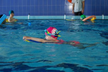 Küçük anaokulu çocuğu havuzda sörf tahtasıyla yüzmeyi öğreniyor. Yüzme dersi. Aktif çocuk suda oynar. Maskeli ve gözlüklü çocuk..
