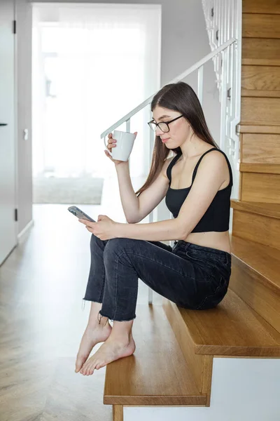 Young woman on stairs at home relaxing, reading email using mobile device and mobile wifi. Cup of hot delicious coffee or tea.