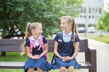 Two children of schoolgirls sit on bench near school, have fun and communicate. Concept of back to school, learning, friendship and childhood