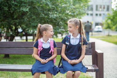 Two children of schoolgirls sit on bench near school, have fun and communicate. Concept of back to school, learning, friendship and childhood