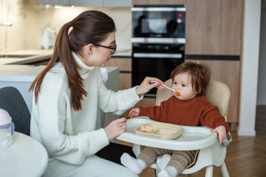 Bebekler için lezzetli balkabağı ve elma püresi. Kahverengi kazaklı küçük kız. Bebek bakıcısı yeni doğmuş küçük bebeği mutfakta besliyor..