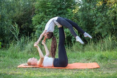 Mother lies on back and keeps daughter on feet during fitness in park. Concept of sports, healthy lifestyle and hobbies