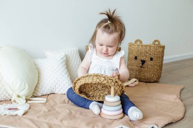 Toddler child plays with developmental toys. Little girl makes pyramid. Basket for toys. Cozy home. Happy baby