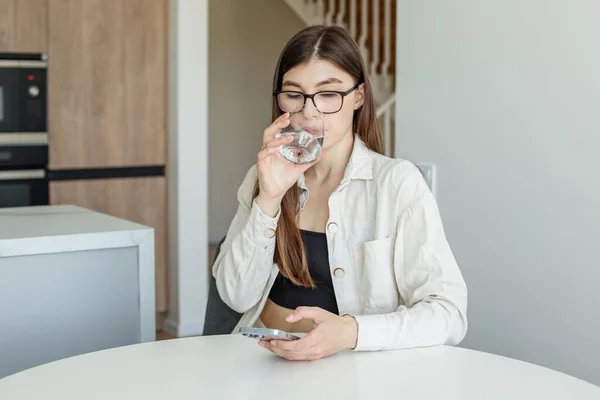 Young adult girl in glasses for vision drinks water and uses mobile phone in home kitchen. Pure water in a glass.