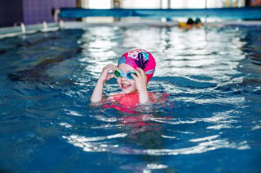 Child schoolgirl learns to swim in pool. Swimming lesson. Active child plays in water. Girl in goggles and swimming cap
