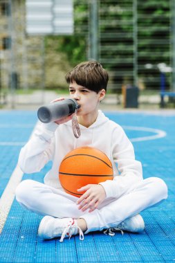 Teenage boy drinks water on basketball court and holds basketball. White tracksuit. Concept of sports, school and active lifestyle