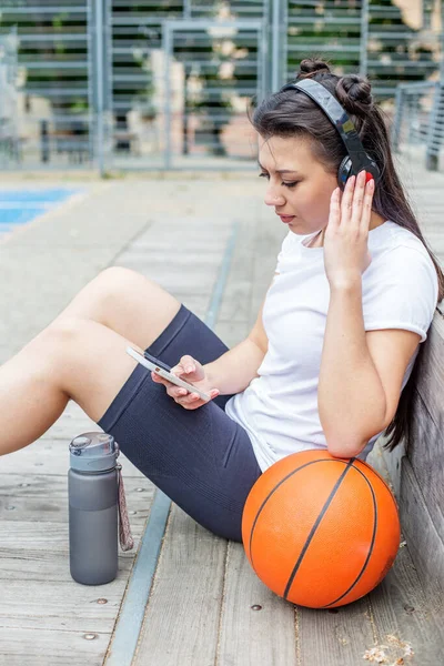 Young adult woman uses wireless headphones and smartphone while sitting on basketball court. Sport, technology and music concept. Reusable water bottle